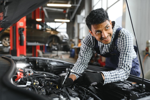 Indischer Automechaniker steht und arbeitet in der Tankstelle Autospezialisten untersuchen das angehobene Auto Professionelle Mechaniker in blauer Mechanikeruniform