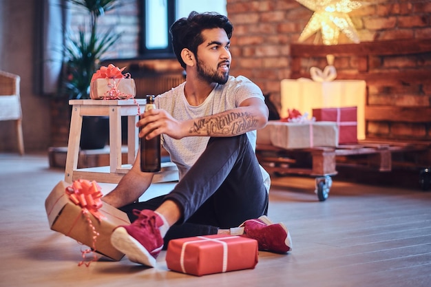 Indischer bärtiger Hipster-Mann mit Tätowierungen auf dem Arm in einem Loft-Zimmer mit Weihnachtsdekoration.