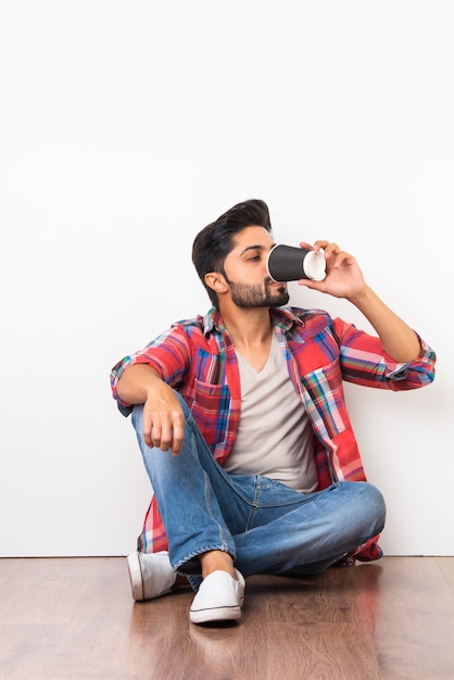 Indischer bärtiger junger Mann trinkt Kaffee aus Einweg-Pappbecher, während er isoliert auf Holzboden vor weißem Hintergrund sitzt