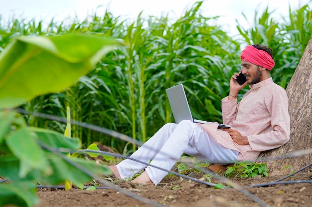 Indischer Bauer, der Laptop benutzt und auf dem Smartphone auf dem Landwirtschaftsfeld spricht.