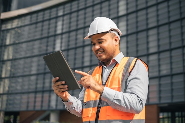 Indischer Bauingenieur des jungen Arbeiters, der Tablette verwendet, um das Projekt zu planen, die moderne Konstruktion zu bauen, die den Bauplan liest Asiatische Architektur oder Ingenieur, der auf der Hochbaustelle arbeitet