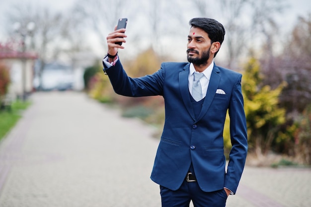 Indischer geschäftsmann mit stylischem bart und bindi auf der stirnabnutzung im blauen anzug posierte im freien und machte selfie am handy