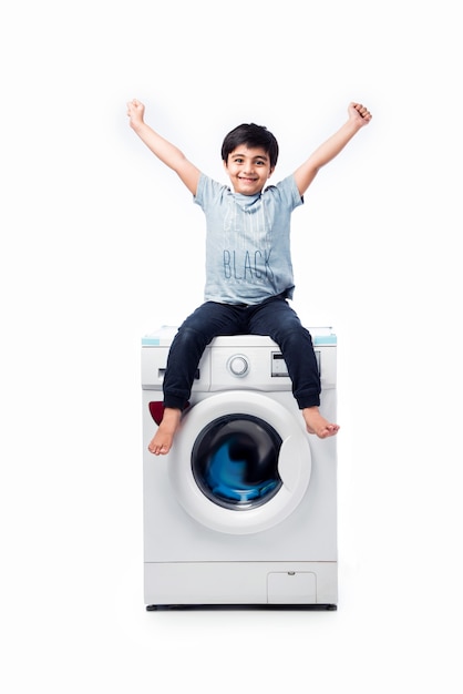 Indischer glücklicher kleiner Junge, der mit Waschmaschine oder Spülmaschine gegen weiße Wand posiert