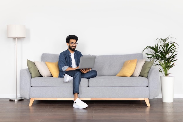 Indischer Mann sitzt auf der Couch und arbeitet zu Hause am Laptop