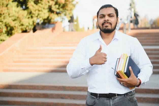 Indischer Student mit Rucksack, der Bücher am sonnigen Tag hält