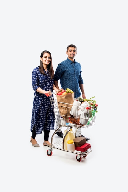 Indisches junges Paar mit Einkaufswagen oder Trolley voller Lebensmittel, Gemüse und Obst. Isoliertes Foto in voller Länge über weißer Wand