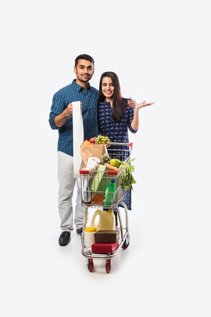 Indisches junges Paar mit Einkaufswagen oder Trolley voller Lebensmittel, Gemüse und Obst. Isoliertes Foto in voller Länge über weißer Wand