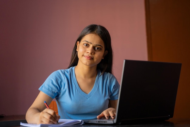 Indisches Mädchen sitzt am Schreibtisch und studiert oder arbeitet am Laptop und macht sich Notizen