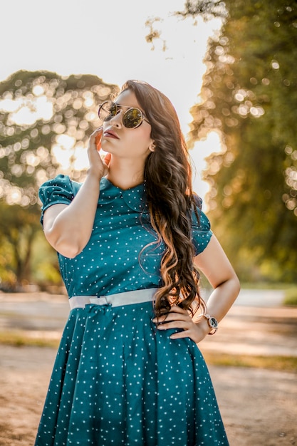 Indisches schönes junges Mädchen mit langen Haaren im Park mit blauem Muster, kurzem Kleid und Sonnenbrille