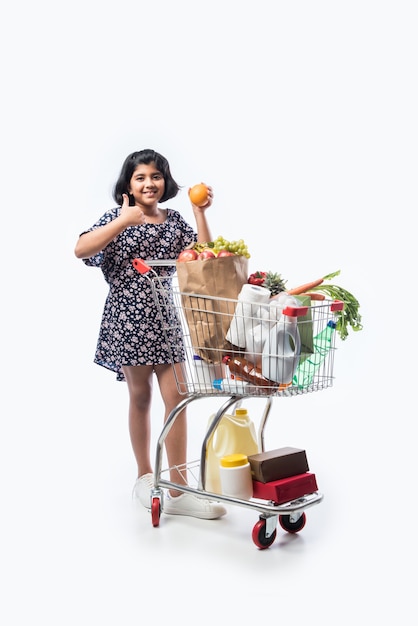 Indisches süßes kleines Mädchen mit Einkaufswagen oder Trolley voll mit Lebensmittelgeschäft, Gemüse und Obst, isoliert über weißer Wand