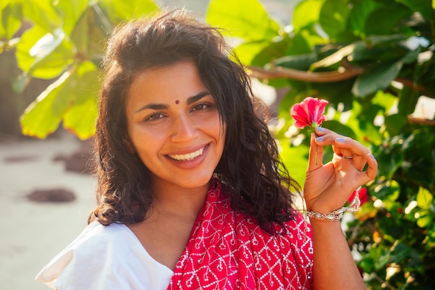 Indisches weibliches Modemodell weißzahniges Lächeln mit Blume in ihrem lockigen Haar im traditionellen indischen Outfit roter Hochzeitssari posiert in der Nähe von tropischen Bäumen an einem paradiesischen Strand Meer ocean.wellness Spa Resort.