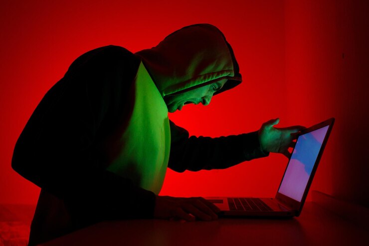 Foto individuo con capucha expresa frustración mientras trabaja en la computadora portátil bajo luz roja