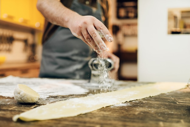 Indivíduo do sexo masculino no avental preparando a massa na mesa da cozinha de madeira. Processo de cozimento de massa caseira