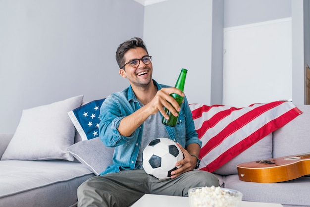 Indivíduo entusiasmado que senta-se em um sofá que come uma cerveja e que olha o futebol.