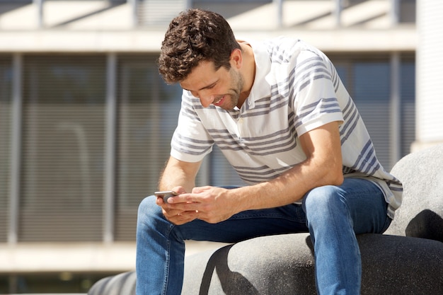 Individuo joven que se sienta al aire libre y que envía el mensaje de texto en el teléfono móvil