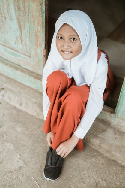 Indonesischer Grundschüler, der sich morgens zur Schule fertig macht und Schuhe vor dem Haus bindet