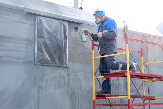 Industriearbeit Grundierung von Metallprodukten aus der Kompressorpistole Ein Arbeiter in Overall und Atemschutzmaske lackiert die Karosserie eines LKW-Anhängers oder eines Metallautos