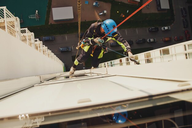 Industrieller Bergsteiger hängt über Wohnfassadengebäude, während er die Außenfassadenverglasung wäscht. Seilzugangsarbeiter hängt an der Hauswand. Konzept der städtischen Werke. Platz für Website kopieren