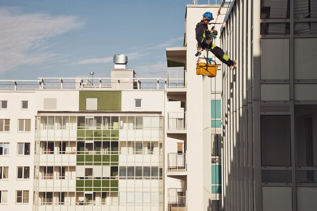 Industrieller Bergsteiger hängt über Wohngebäude, während er die Außenfassadenverglasung wäscht. Seilzugangsarbeiter hängt an der Hauswand. Konzept der städtischen Werke. Platz kopieren