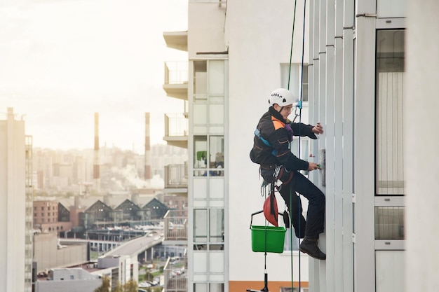 Industrieller Bergsteiger in Uniform hängt über Wohngebäude im Hintergrund der Stadt und wäscht Außenfassade Seilzugangsarbeiter an der Hauswand