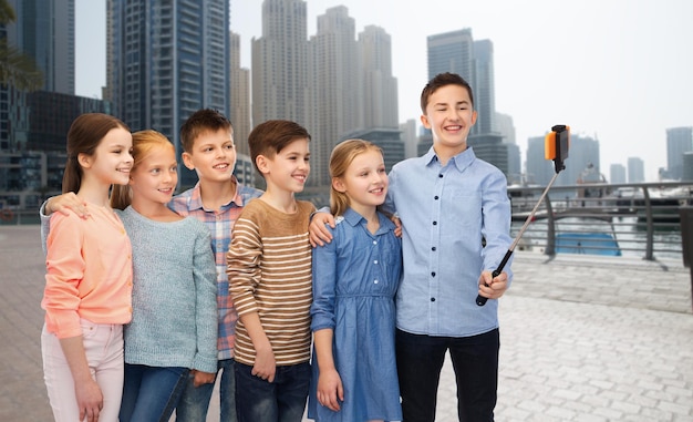 Infancia, amistad, tecnología y concepto de personas - grupo de niños felices hablando de imágenes por teléfono inteligente en selfie stick sobre el fondo de la calle de la ciudad de dubai