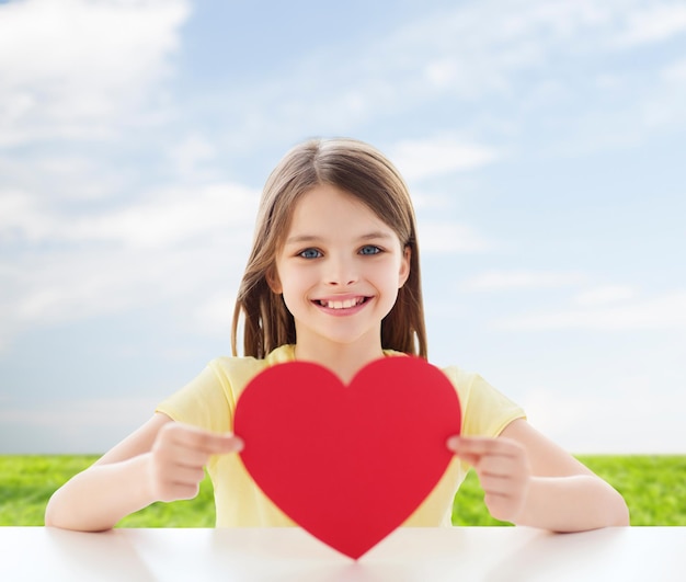 la infancia, el amor, la caridad, el medio ambiente y el concepto de la gente - una niña sonriente sentada y sosteniendo un corazón rojo recortado sobre el fondo natural