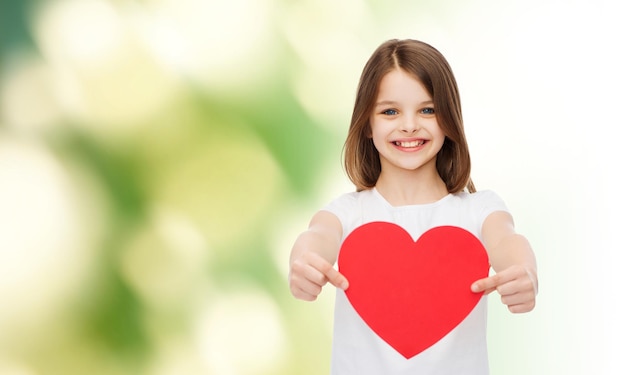 infância, amor, caridade, ecologia e conceito de pessoas - menina sorridente sentada e segurando o recorte de coração vermelho sobre fundo verde