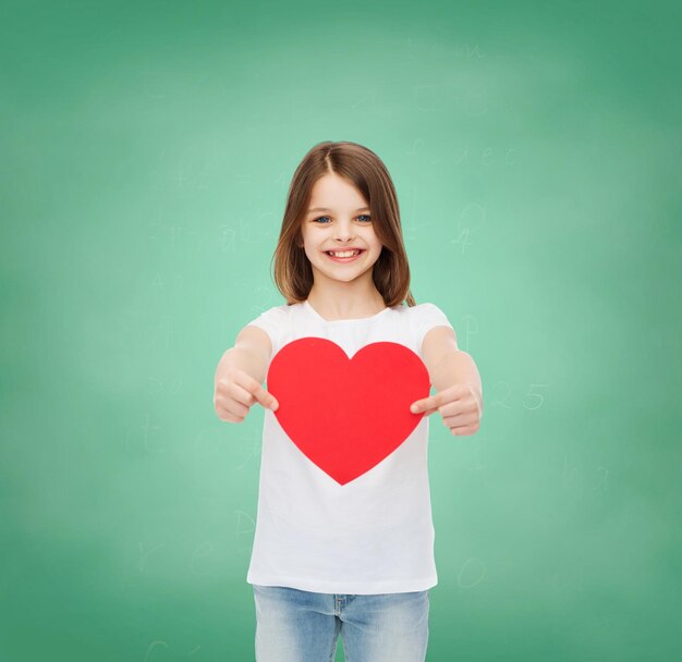 infancia, caridad, educación, amor y concepto de la gente - niñita sonriente sentada con un corte de corazón rojo sobre fondo de pizarra verde