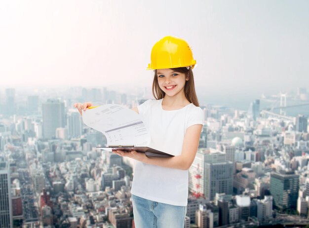 Infancia, construcción, arquitectura, construcción y concepto de personas - niña sonriente con casco protector con página de giro del portapapeles