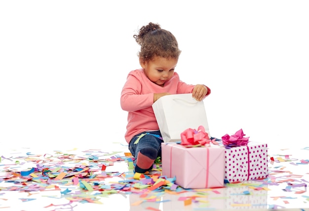 Infancia, cumpleaños, fiesta, vacaciones y concepto de la gente - feliz y sonriente niña afroamericana con cajas de regalo y confetti jugando con una bolsa de compras sentada en el suelo