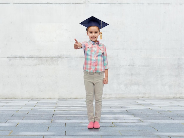 la infancia, la escuela, la educación, el aprendizaje y el concepto de la gente - niña feliz con sombrero de soltero o mortarboard mostrando los pulgares hacia arriba sobre el fondo de hormigón urbano