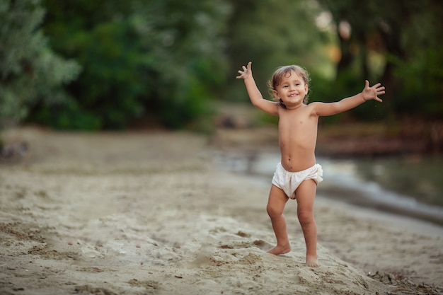 Infancia feliz alegre niño emocional niña 1-2 años juega en la orilla arenosa cerca del río. árboles verdes crecen detrás de ella