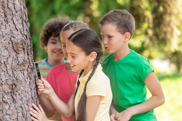 Infancia feliz. Chicas y chicos interesados en mirar con lupa cerca del árbol en el parque en verano