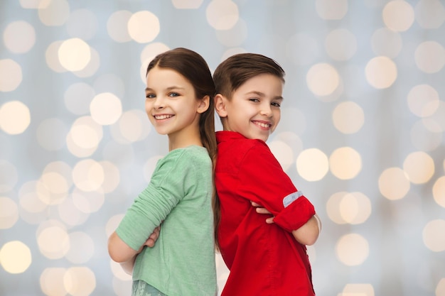 Foto infancia, moda y concepto de la gente - niño y niña sonrientes felices de pie espalda con espalda durante las vacaciones luces de fondo