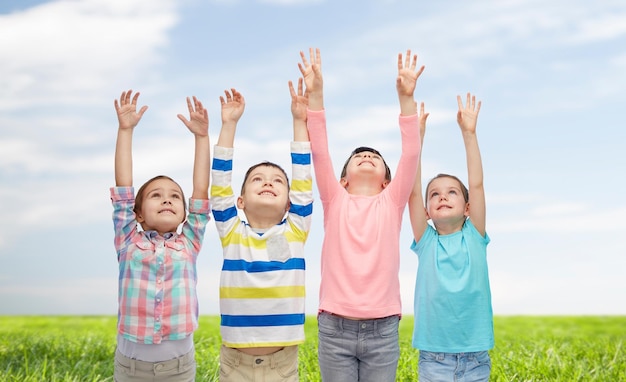 infancia, moda, gesto y concepto de la gente - niños sonrientes felices levantando la mano y celebrando la victoria sobre el cielo azul y el fondo de la hierba