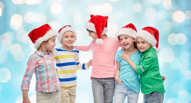 infância, natal, feriados, amizade e conceito de pessoas - grupo de crianças sorridentes felizes em chapéus de papai noel abraçando sobre o fundo azul das luzes de férias