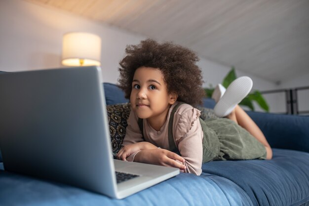 Infância, passatempo. Menina afro-americana pensativa deitada no sofá em casa na frente do laptop