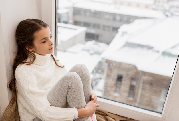 Foto infancia, tristeza y concepto de la gente - triste niña hermosa en suéter sentada en el alféizar de la ventana de la casa en invierno