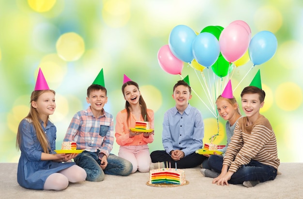 Infancia, vacaciones, celebración, amistad y concepto de la gente: niños sonrientes felices con sombreros de fiesta con pastel de cumpleaños y globos sobre el fondo verde de las luces de las vacaciones de verano