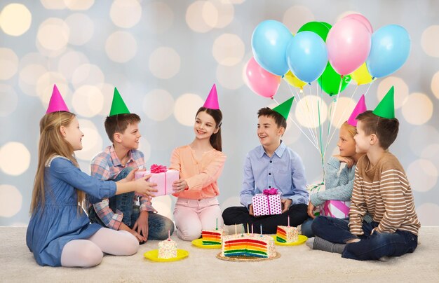 infancia, vacaciones, celebración, amistad y concepto de la gente - niños sonrientes felices con sombreros de fiesta con pastel dando regalos en la fiesta de cumpleaños sobre fondo de luces