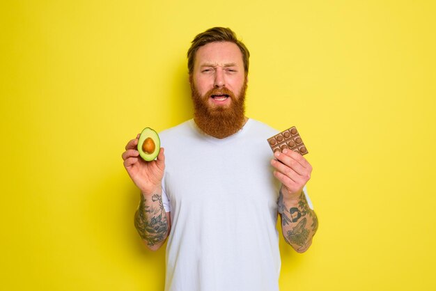 Infeliz hombre con barba y tatuajes tiene aguacate y chocolate