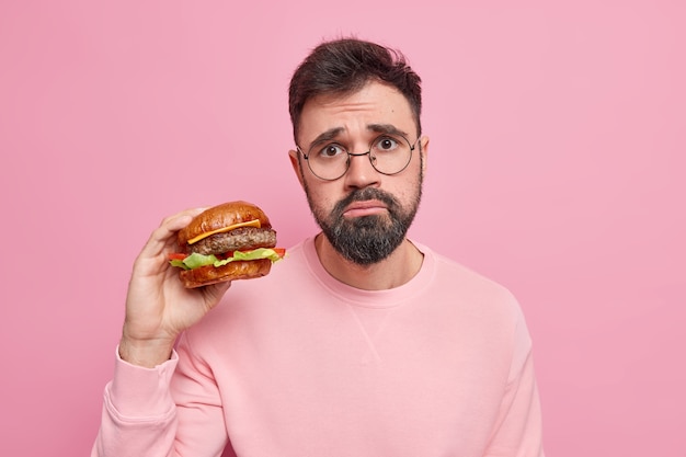 Infeliz hombre europeo barbudo y hambriento obtiene un refrigerio alto en calorías sostiene apetitosa hamburguesa come comida poco saludable usa gafas redondas y suéter