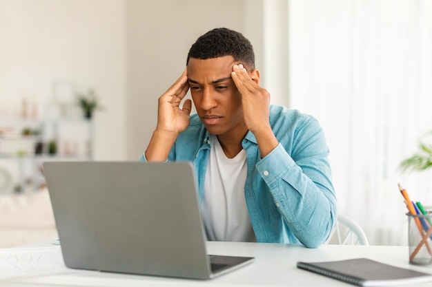Infeliz joven afroamericano mira la computadora que sufre de estrés y migraña en el interior de la habitación o la oficina