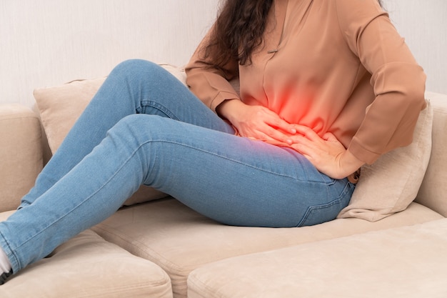 Infeliz mujer asiática sentada en el sofá y sosteniendo el sufrimiento del estómago. Dolor abdominal que proviene de la menstruación, diarrea o indigestión. Concepto de salud y enfermedad