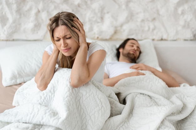 Infeliz mujer enojada sentada en la cama y cubriendo sus oídos con las manos que sufren de los ronquidos de su marido