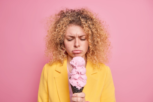 La infeliz mujer europea de pelo rizado mira con tristeza el helado siente que la tentación no puede permitirse comer alimentos ricos en calorías vestida con ropa amarilla aislada sobre una pared rosa. Helado sabroso