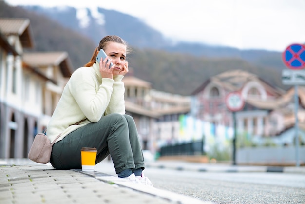 Infeliz mujer triste habla por un teléfono celular niña sentada en la acera al lado de la carretera hablando por un teléfono inteligente