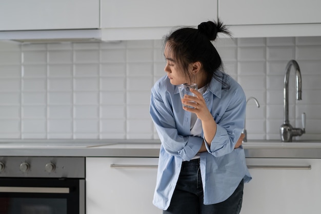 Infeliz mulher asiática sofrendo com problemas digestivos em pé na cozinha com um copo de água