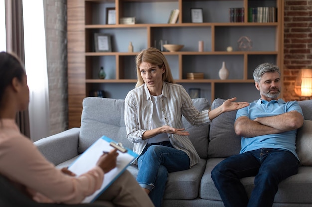 Infeliz mulher europeia de meia-idade mostra para homem ofendido no sofá no interior do escritório do psicólogo