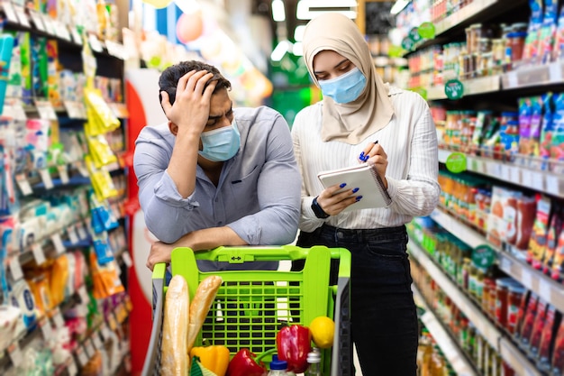 Infeliz pareja de familia musulmana calculando los precios de las compras en el supermercado luchando contra la crisis financiera. Marido árabe cansado esperando a la esposa mientras toma notas en la lista de la tienda